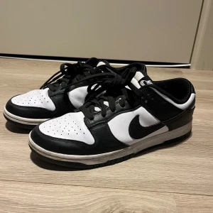 Nike Dunk Low svartvita sneakers - Säljer ett par Nike Dunk Low sneakers i svart och vitt. Skorna har klassisk låg modell med svart swoosh, snörning och perforerad tå. Perfekta för dig som gillar streetwear och vill ha en stilren look. Har tyvärr inte kvar originalskolådan men skorna köptes för ca 2 års sedan och har använts till och från. Är i fint skick. 