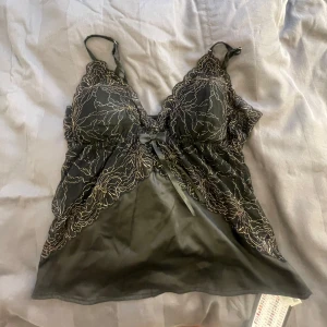 Svart spetslinne med rosett - Säljer ett svart linne med tunna axelband och vacker spets upptill. Linnet har en liten rosett framtill och transparent tyg med broderade detaljer. Helt ny, köpte fel storlek, mindre i storleken men storlek XL