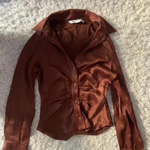 Brun satinblus från Zara - Snygg brun långärmad blus från Zara i glansigt satinmaterial. Skjortan har klassisk krage, knappar framtill och lätt rynkade detaljer vid midjan för en smickrande passform. Perfekt att styla till olika looks.