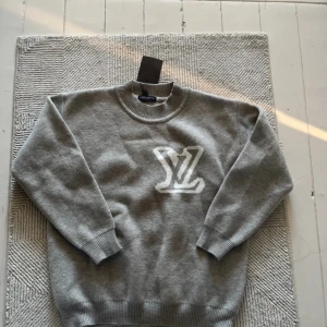 Grå stickad tröja från Louis Vuitton - Säljer en grå stickad tröja från Louis Vuitton med stor vit LV-logga på bröstet. Tröjan har rund halsringning och ribbade muddar vid ärmslut och nederkant. Perfekt för dig som gillar exklusiva märken och stilren design.