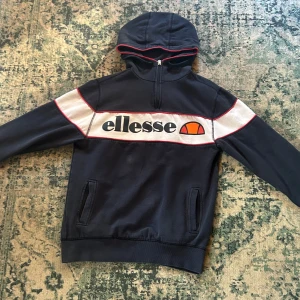 Mörkblå hoodie från Ellesse med logga - Säljer en mörkblå hoodie från Ellesse med vit och röd detalj över bröstet och stor logga. Tröjan har huva, dragkedja vid halsen och fickor framtill. Perfekt för en sportig och avslappnad stil.