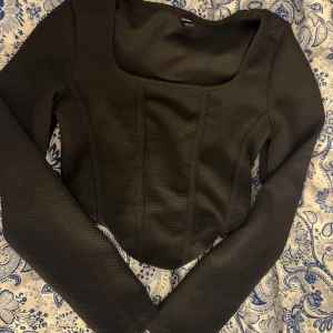 Svart långärmad topp med fyrkantig ringning - Säljer en svart långärmad topp från Bershka med fyrkantig ringning och markerade sömmar framtill. Toppen har en figurnära passform och är tillverkad i ett strukturerat material som ger en snygg siluett.