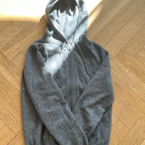 Grå hoodie i kashmir från Soft Goat - Mjuk och lyxig grå hoodie från Soft Goat i 100% kashmir. Tröjan har dragkedja framtill, huva med dragsko och två fickor. Perfekt för en avslappnad och stilren look. Väldigt grisch.