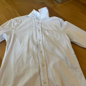 Vit och blå randig skjorta från Zara - Säljer en klassisk vit skjorta med tunna blå ränder från Zara. Skjortan har lång ärm, bröstficka och knäppning framtill. Perfekt för en stilren look och enkel att matcha med jeans eller kostymbyxor.