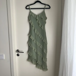 Grön klänning  - Säljer en ljusgrön klänning med smala axelband och volangdetaljer som går diagonalt över hela klänningen. Klänningen har en asymmetrisk nederkant och är tillverkad i ett lätt, flowy material som ger en somrig känsla.