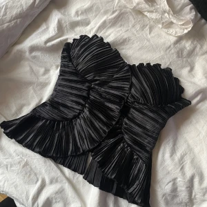 Svart plisserad tubtopp - Säljer en svart plisserad tubtopp med volangdetaljer. Toppen är axelbandslös och har en snygg struktur som ger en unik look. Perfekt för dig som vill sticka ut med en trendig och elegant topp.