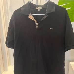 Svart pikétröja från Burberry - Klassisk svart pikétröja från Burberry med korta ärmar och den ikoniska rutiga detaljen vid kragen. Liten broderad logga på bröstet. Perfekt för en stilren och avslappnad look. Passar lite större, så funkar definitivt för size L