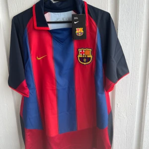 FC Barcelona fotbollströja Ronaldinho #10 - Klassisk FC Barcelona fotbollströja med Ronaldinho och nummer 10 på ryggen. Tröjan är randig i rött och blått med gula detaljer, klubbmärke på bröstet och LFP-märke på ärmen. Perfekt för fans av laget och legenden.