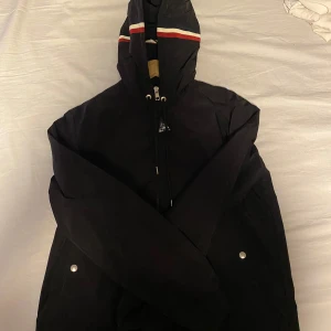 Svart vindjacka med huva och ränder - Svart vindjacka med huva och dragkedja framtill. Huvan har röda och vita ränder som detalj. Jackan har två fickor med tryckknappar och justerbar dragsko vid huvan. Perfekt för blåsiga dagar.