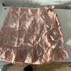 Blommig omlottkjol med volang och knyt - Supersöt kort omlottkjol med blommigt mönster i pastelliga färger. Kjolen har volang längs nederkanten och knyts i midjan. Perfekt för en somrig outfit och lätt att matcha med olika toppar.💕☀️⛱️🍉🌺