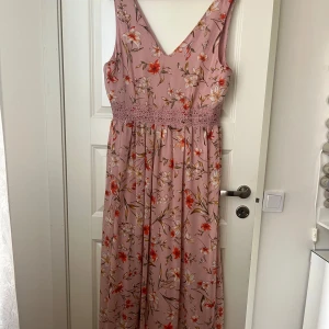 Ljusrosa blommig långklänning med spetsdetalj - Säljer en romantisk långklänning i ljusrosa med blommigt mönster och v-ringning. Klänningen har breda axelband och en vacker spetsdetalj i midjan. Perfekt för sommarens tillfällen!