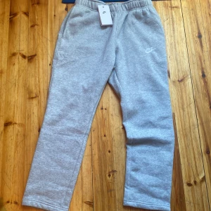 Grå mjukisbyxor från Nike - Säljer ett par klassiska grå mjukisbyxor från Nike med vit logga på vänster lår. Byxorna har resår i midjan och raka ben, samt sidofickor. Perfekta för chill eller träning.