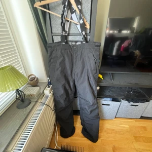 Svarta täckbyxor med hängslen från Ski Industries - Svarta täckbyxor från Ski Industries med justerbara hängslen och tryckknapp i midjan. Byxorna är vindtäta och vattenresistenta, perfekta för kalla dagar. Klassisk passform och diskret design med logga på hängslena.