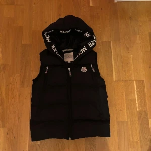 Svart dunväst med huva från Moncler - Säljer en svart dunväst från Moncler med huva och dragkedja framtill. Västen har två fickor med dragkedja och Moncler-logga på bröstet. Insidan av huvan har Moncler-tryck. Perfekt för lager-på-lager-stil.