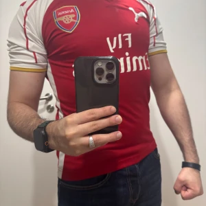Arsenal fotbollströja Puma - Röd Arsenal fotbollströja från Puma med vita ärmar och gula detaljer. Klassisk klubbsköld på bröstet och Puma-logga. Kortärmad modell med sportig passform, perfekt för match eller träning.