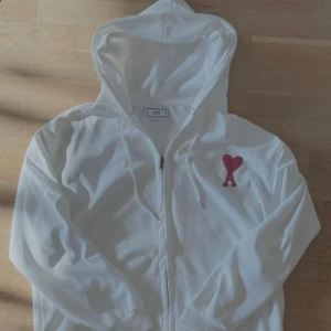 Vit hoodie med rött hjärta från Ami - Säljer en vit hoodie från Ami med dragkedja och huva. På bröstet finns ett broderat rött hjärta med bokstaven A. Perfekt för en stilren och avslappnad look.