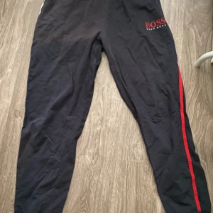 Mörkblå mjukisbyxor från Hugo Boss med röd logga - Mörkblå mjukisbyxor från Hugo Boss med logga på ena benet och röd rand längs sidorna. Byxorna har snörning i midjan och är tillverkade i mjukt material. Perfekta för en avslappnad stil.