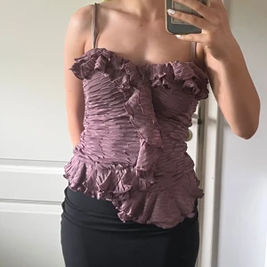 Ljuslila volang topp med smala axelband - Säljer en ljuslila topp med smala axelband och volangdetaljer. Toppen har en plisserad struktur och asymmetriska volanger både framtill och nertill. Perfekt för dig som gillar unika detaljer och en feminin stil.