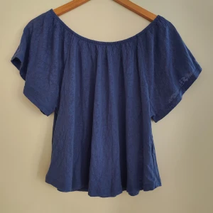Blå offshoulder topp från Gina Tricot - Säljer en blå offshoulder topp från Gina Tricot. Toppen har korta ärmar och är tillverkad i ett lätt och luftigt material, perfekt för varma dagar. Enkel och stilren design med blommigt mönster som passar till mycket.
