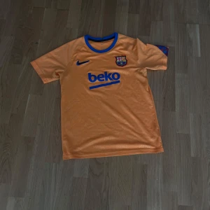 Barcelona orange fotbollströja från Nike - Säljer en orange FC Barcelona fotbollströja från Nike med blå detaljer och klubbmärke på bröstet. Tröjan har korta ärmar, blå krage och BEKO sponsortryck på framsidan. Perfekt för dig som älskar Barca!