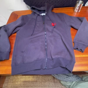 Mörkblå hoodie med dragkedja från Comme des Garçons Play - Mörkblå hoodie med dragkedja och huva från Comme des Garçons Play. Klassisk modell med fickor framtill och röd hjärtlogga på bröstet. Perfekt för en avslappnad stil. Pris kan diskuteras lite.