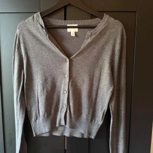 Grå kofta från H&M - En enkel och stilren grå kofta från H&M med långa ärmar och knäppning framtill. Tröjan har en klassisk passform och ribbade muddar vid ärmslut och nederkant. Perfekt att slänga på sig över en topp.