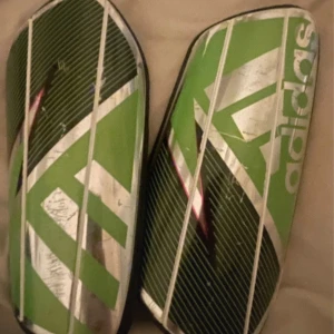 Gröna benskydd från Adidas - Säljer ett par gröna och svarta benskydd från Adidas med vitt mönster och logga. De har en böjd form för att sitta bra på benet och är tillverkade i hård plast med mjuk baksida för komfort.