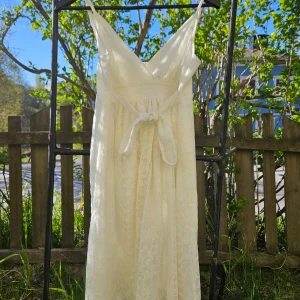 Ljus långklänning med knytband - Romantisk ljus långklänning med v-ringad hals. Klänningen har ett fint knytband i midjan och är tillverkad i ett lätt, spetsmaterial som ger en elegant känsla.