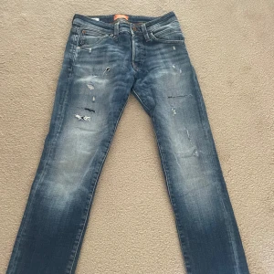 Jack & Jones jeans med slitningar - Säljer ett par blå slim fit jeans, modell Glenn. Jeansen har slitna detaljer framtill och klassisk femficksdesign. De har en något tvättad look och passar perfekt till en avslappnad stil. Modellen är 28/30 priset är 299, ej hugget i sten.