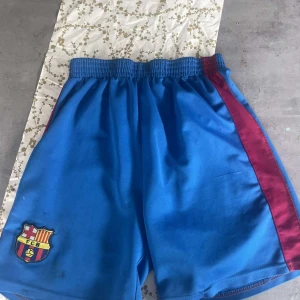 Blå FC Barcelona shorts med röd detalj - Säljer ett par blå shorts från FC Barcelona med klubbens emblem på vänster ben och en röd rand längs sidan. Perfekta för fotboll eller träning. Resår i midjan för skön passform.