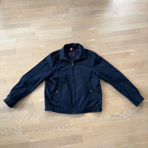 Marinblå vindjacka från Tommy Hilfiger - Snygg marinblå tunn vår/sommarjacka från Tommy Hilfiger med dragkedja framtill och två fickor med knappar. Klassisk krage och diskret logga på bröstet. Perfekt för svala dagar och enkel att matcha med olika outfits.