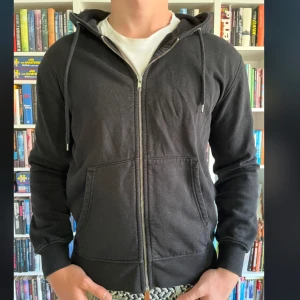 Svart hoodie med dragkedja från Colorful Standard - Säljer en svart hoodie med dragkedja från Colorful Standard. Hoodien har huva med snörning, två fickor framtill och ribbade muddar vid ärmslut och nederkant. Perfekt för en avslappnad stil. Nypris är 1000kr 