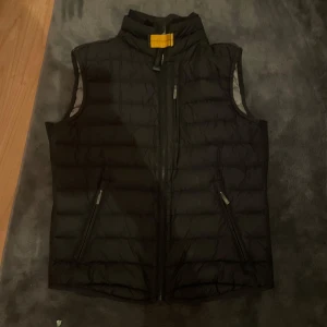 Svart dunväst från Parajumpers - Svart dunväst från Parajumpers med hög krage och dragkedja framtill. Västen har två snedställda fickor med dragkedja och ett broderat märke på ryggen vid nacken. Perfekt för lager-på-lager och stilren look. Storleken är 24-26. Finns inga skador eller någonting på den heller och finns qr så man kan checka om den är riktig