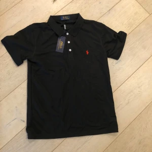 Svart pikétröja från Polo Ralph Lauren - Säljer en klassisk svart pikétröja från Polo Ralph Lauren med röd broderad logga på bröstet. Tröjan har korta ärmar, krage och knappar framtill. Perfekt för en stilren och avslappnad look. Perfekt inför sommaren✅🤩