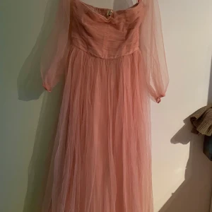 Rosa långklänning från Lace & Beads - Romantisk långklänning i dusty pink från Lace & Beads. Klänningen har transparenta långa ärmar och ett luftigt, voluminöst tyg i flera lager. Perfekt för speciella tillfällen när du vill känna dig extra fin.