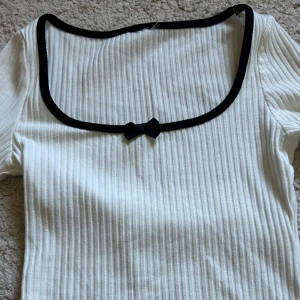 Vit ribbad topp med svart kant och rosett - Säljer en vit ribbad topp med lång ärm. Toppen har en fyrkantig urringning med svart kant och en liten svart rosett framtill. Ärmsluten och nederkanten har svarta vågiga detaljer. Perfekt för dig som gillar stilrena detaljer.