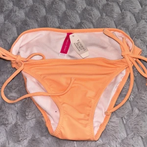 Persikofärgad bikiniunderdel från Victoria's Secret - Säljer en persikofärgad bikiniunderdel från Victoria's Secret med knyt i sidorna. Bikinin har en enkel och stilren design med tunna band och är tillverkad i mjukt material som känns bekvämt mot huden.