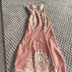 Rosa bal/fest långklänning med broderade blommor - Säljer en elegant rosa långklänning med tunna axelband och vacker vit broderad blommönster framtill och baktill. Klänningen har figurnära passform och dekorativa pärlor längs ryggen. Perfekt för speciella tillfällen!