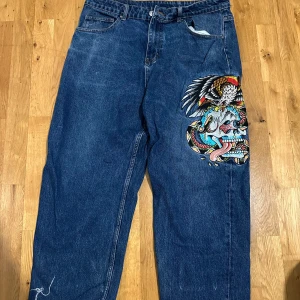 Blå jeans med drakmotiv från Ed Hardy - Unika blå jeans från Ed Hardy med stort färgglatt drakmotiv på ena benet och broderad logga på bakfickan. Modellen är loose fit med råa kanter nertill för en cool streetstil.