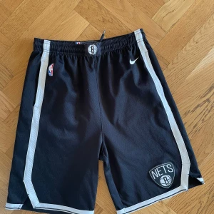 Brooklyn Nets basket shorts - Säljer ett par svarta Brooklyn Nets shorts från Nike med vita detaljer och elastisk midja. Perfekta för basket eller avslappnad stil. NBA-logga och Nets-emblem på benen.