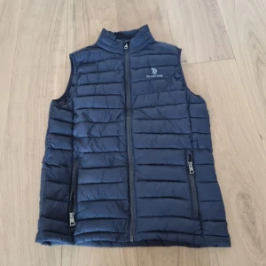 Blå dunväst från U.S. Polo Assn. - Snygg blå dunväst från U.S. Polo Assn. med dragkedja framtill och två praktiska fickor med dragkedjor. Västen har ett stilrent broderat märke på bröstet och är perfekt för kyligare dagar. 