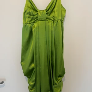 Grön axelbandslös klänning - Snygg grön axelbandslös klänning i satin med draperad design. Perfekt för en elegant look med en unik stil. Klänningen har en vacker rosettdetalj framtill som ger en extra touch.