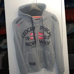 Grå hoodie från Geographical Norway - Säljer en riktigt snygg å skön  hoodie från Geographical Norway med dragkedja och tryck av norska flaggan på framsidan. Tröjan har en justerbar huva med dragsko och två fickor framtill. Tröjan är helt ny och aldrig använd, strl S. Nypris ca 1200, mitt pris 500!