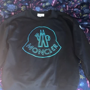 Svart sweatshirt från Moncler - Säljer en svart sweatshirt från Moncler med ett stort turkost tryck på framsidan. Tröjan har en rund halsringning och långa ärmar. Perfekt för en stilren och avslappnad look.
