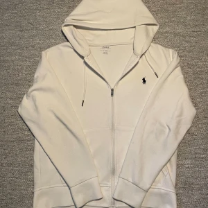 Vit hoodie från Polo Ralph Lauren - Säljer en stilren vit hoodie från Polo Ralph Lauren med dragkedja och klassisk logga på bröstet. Perfekt för en avslappnad look. Den har en justerbar huva med snören och långa ärmar.