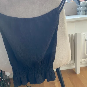 Svart topp - Snygg svart topp med smala axelband och volangdetalj i nederkant. Perfekt för en stilren look. Skicka gärna prisförslag