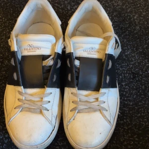 Valentino Garavani sneakers - Snygga vita Valentino Garavani sneakers med svart banddetalj över snörningen. Skorna har en rund tå och klassisk snörning. Perfekta för en stilren look.