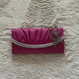 Rosa kuvertväska med kedja - Snygg rosa kuvertväska med silverdetaljer och kedja. Väskan har en elegant rynkad design framtill och en praktisk insida med flera fack. Perfekt för att bära med sig det viktigaste på en utekväll.