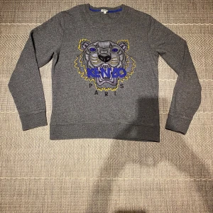 Grå sweatshirt från Kenzo - Säljer en grå sweatshirt från Kenzo med ett broderat tigerhuvud och Kenzo-logga i blått och gult på framsidan. Säljer denna eftersom att den är för stor för mig. Skick: 8/10