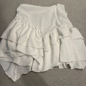 Vit volangkjol - Säljer en söt vit kortkjol med volanger. Kjolen har en omlott-design som ger en fin rörelse och stil. Perfekt för varma dagar och somriga tillfällen. Pris kan diskuteras och jag är villig till att sänka priset!!!